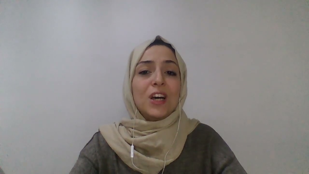 A woman wearing a beige hijab and a dark top is speaking. She has a wired earbud in her left ear and is seated in front of a plain white wall.