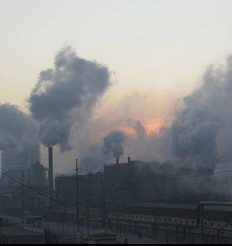 A factory complex emits large clouds of smoke from multiple smokestacks, obscuring the sky as the sun sets in the background. Power lines are visible in the foreground.