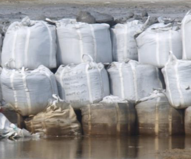 Large white industrial sacks, some stained and weathered, are stacked in two rows beside muddy water, with more sacks and a barren landscape visible in the background.