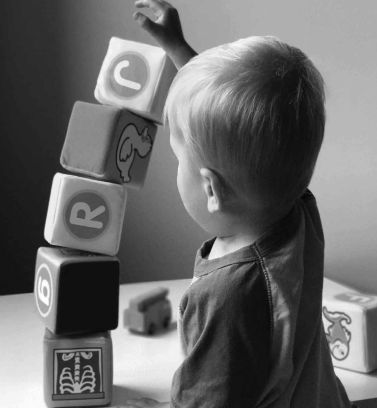 A young child with light hair, seen from behind, reaches up to stack large toy blocks on a table. The blocks display various letters and images. The photo is in black and white.
