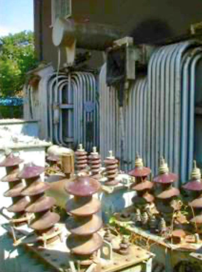 An outdoor electrical substation with several large ceramic insulators, metal structures, and coiled pipes mounted on a building wall. The area is exposed to sunlight and partially shaded.