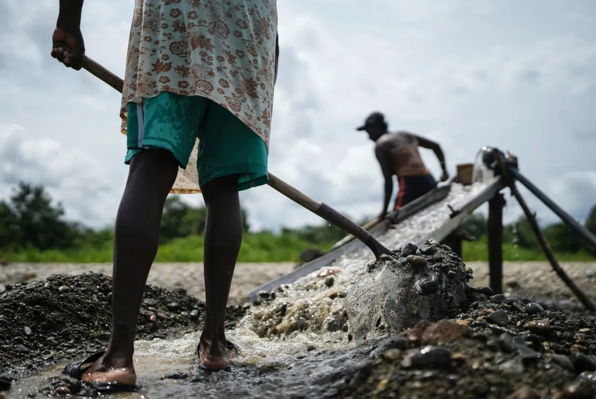 Two people work outdoors using tools to sift and wash gravel in shallow water. One person stands close, seen from behind holding a shovel, while the other is further away operating a sluice. The sky is cloudy.