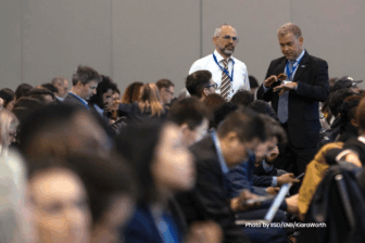 A large group of people sit in rows at a conference or meeting. Two men in business attire stand in the aisle, engaged in conversation. Most attendees appear focused on their devices or notes.