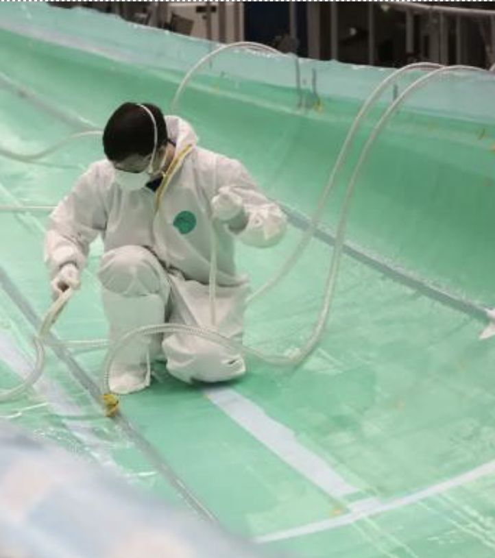 A person wearing a white protective suit and face mask is kneeling inside a large, empty, turquoise industrial tank while handling hoses.