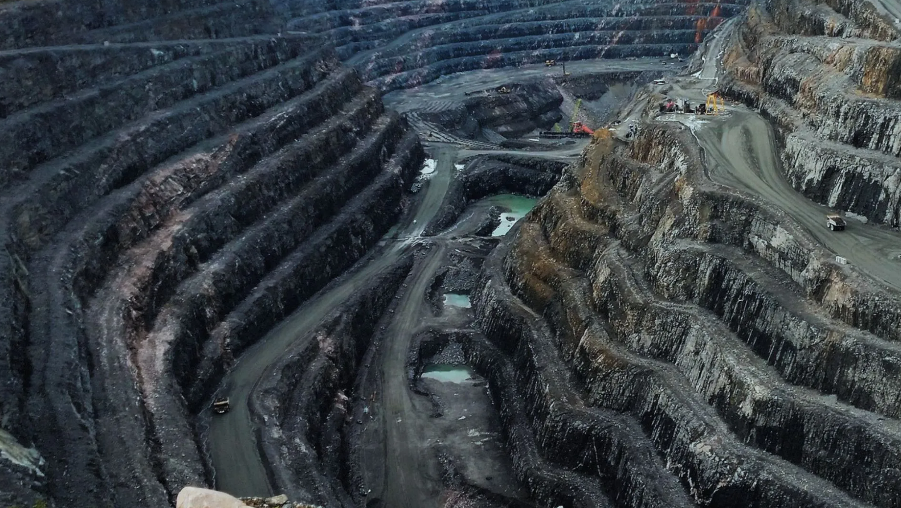 A large open-pit mine with stepped, terraced sides, winding roads, and several pools of water at the bottom. Machinery and vehicles are visible on different levels of the excavation.