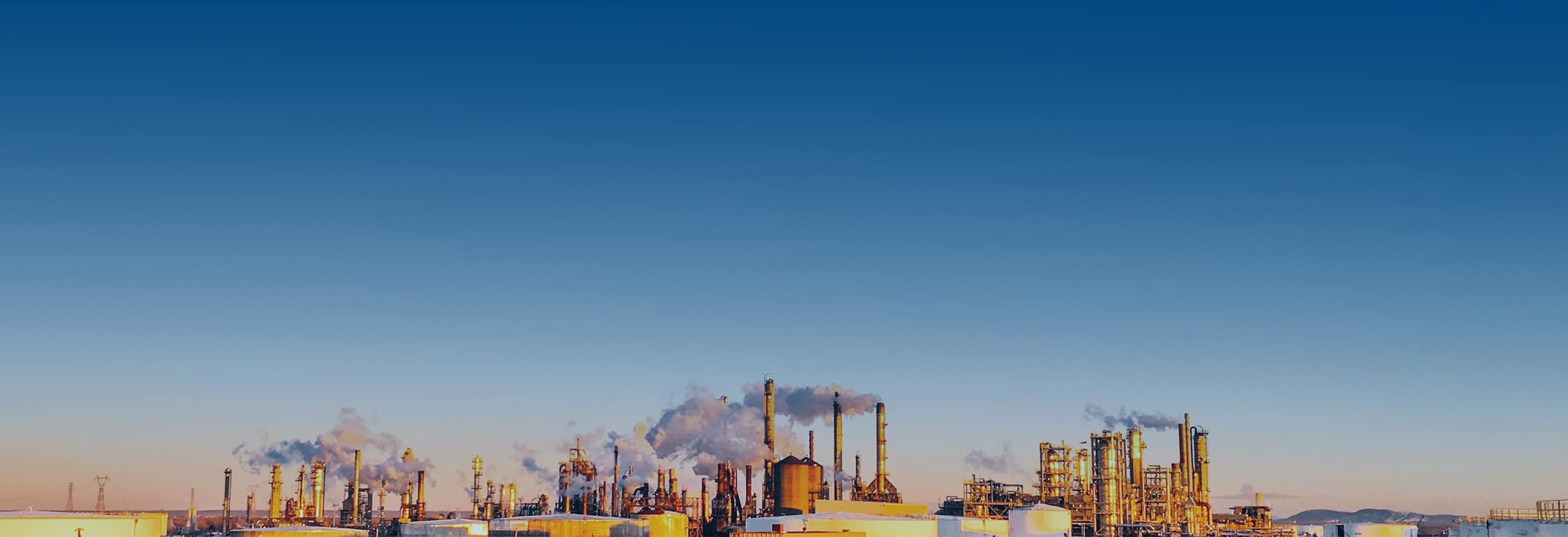 An industrial refinery complex with numerous smokestacks emitting smoke under a clear blue sky, with low buildings and equipment spread across the foreground.