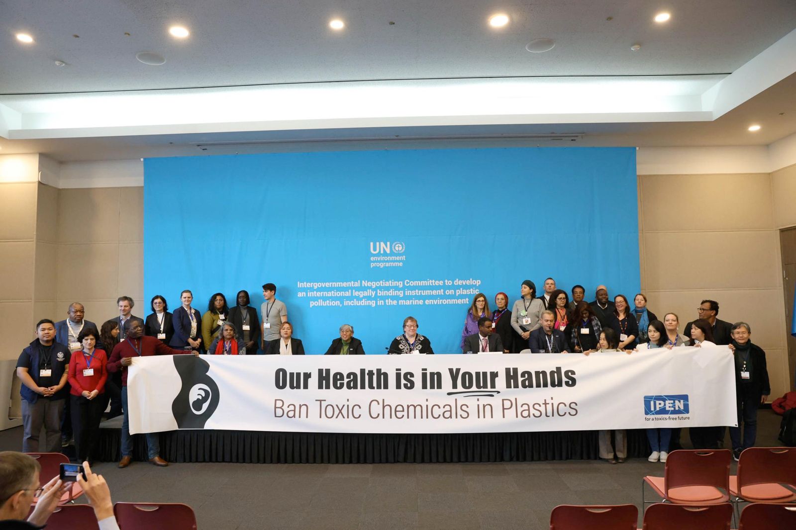 A large group of people stand on a stage holding a banner that reads, Our Health is in Your Hands. Ban Toxic Chemicals in Plastics. Behind them is a blue sign displaying the UN logo and text about plastic pollution.