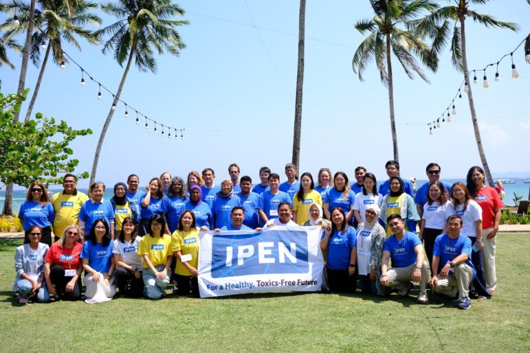 A large group of people pose outdoors on grass under palm trees, holding a banner that reads IPEN For a Healthy, Toxics-Free Future. Most are wearing blue or yellow IPEN shirts; the ocean and clear sky are visible in the background.