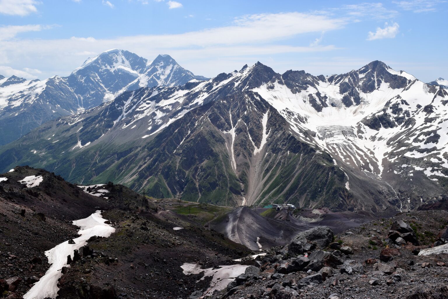 Snow-capped mountains with rugged peaks and valleys are seen under a partly cloudy sky. Patches of snow and rocky terrain are visible in the foreground, with a mix of green areas on lower slopes.