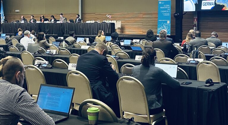 A conference room with attendees seated at tables using laptops, facing a panel of speakers at the front. Name signs for countries are visible, and a large screen displays a blue graphic with text.