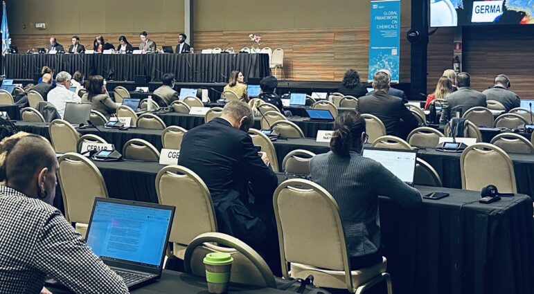 A conference room with attendees seated at tables using laptops, facing a panel of speakers at the front. Name signs for countries are visible, and a large screen displays a blue graphic with text.