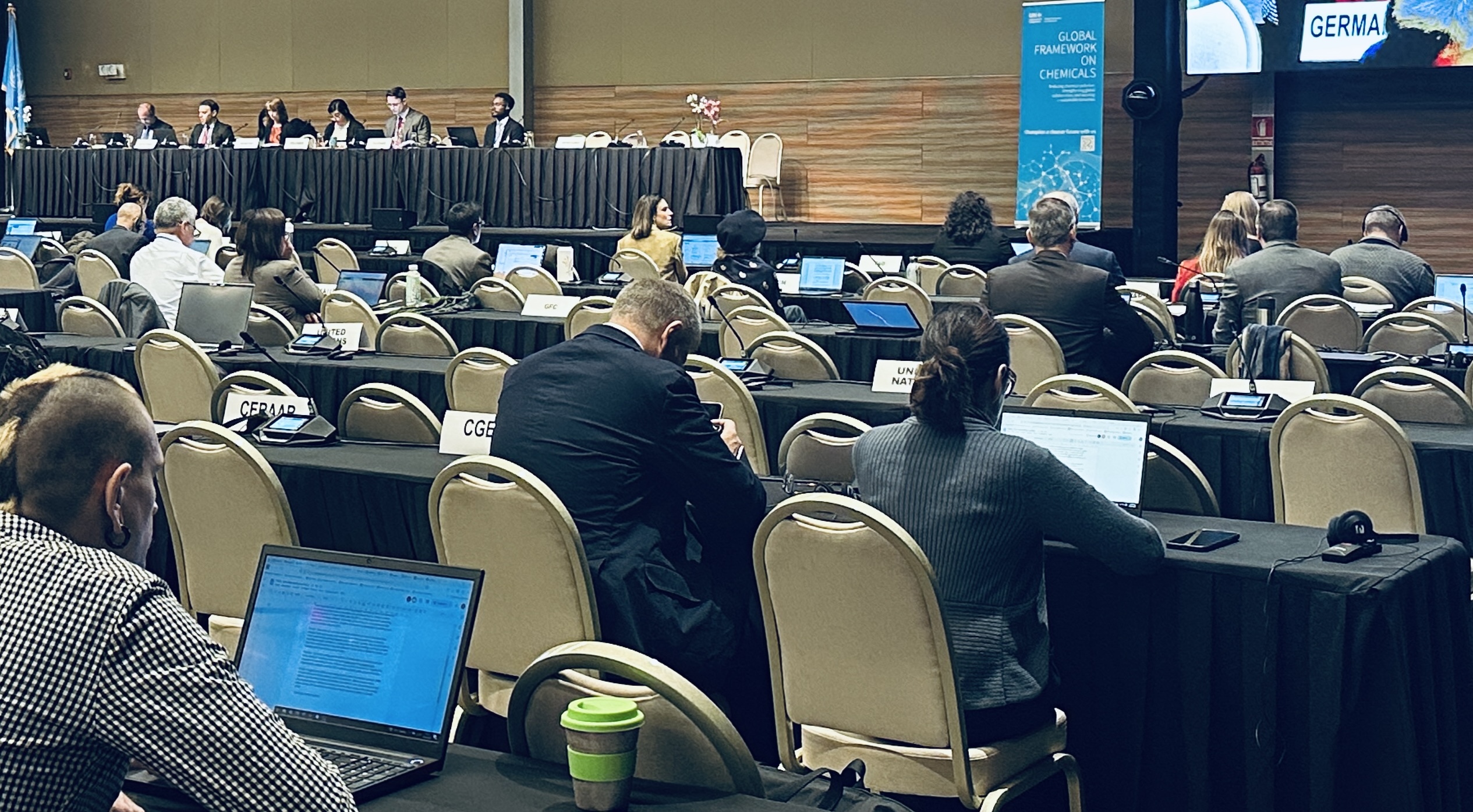 Uma sala de conferências com participantes sentados em mesas usando laptops, de frente para um painel de palestrantes na frente. Placas com os nomes de países são visíveis, e uma grande tela exibe um gráfico azul com texto.
