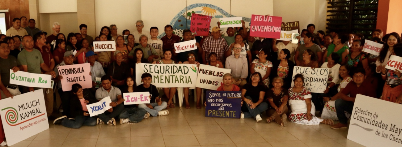 A large group of people stand indoors holding colorful signs with various messages, including “Seguridad Alimentaria,” “Pueblos Unidos,” and “Quintana Roo,” with a mural in the background.