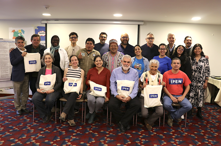 A group of 20 people pose indoors for a photo, some sitting and some standing, with several holding white tote bags labeled IPEN. The background shows charts and a map on the wall. The carpet is red with a blue and white pattern.