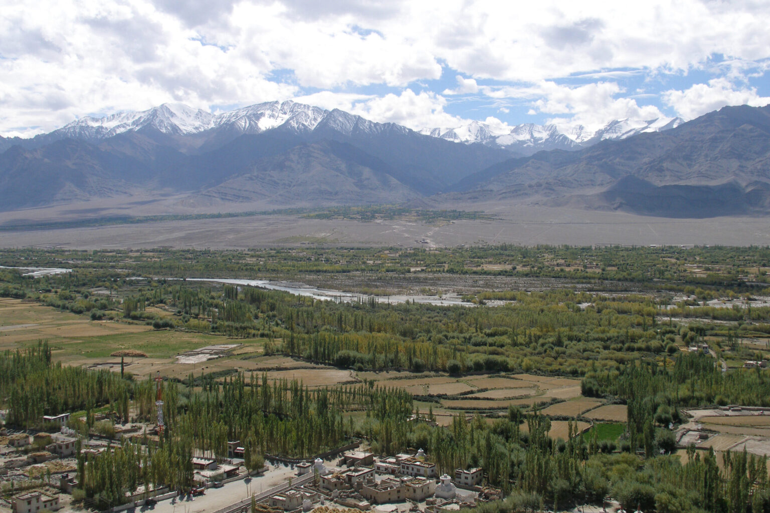 A wide valley with green fields and scattered trees in the foreground, a river running through the middle, and snow-capped mountains under a partly cloudy sky in the background.