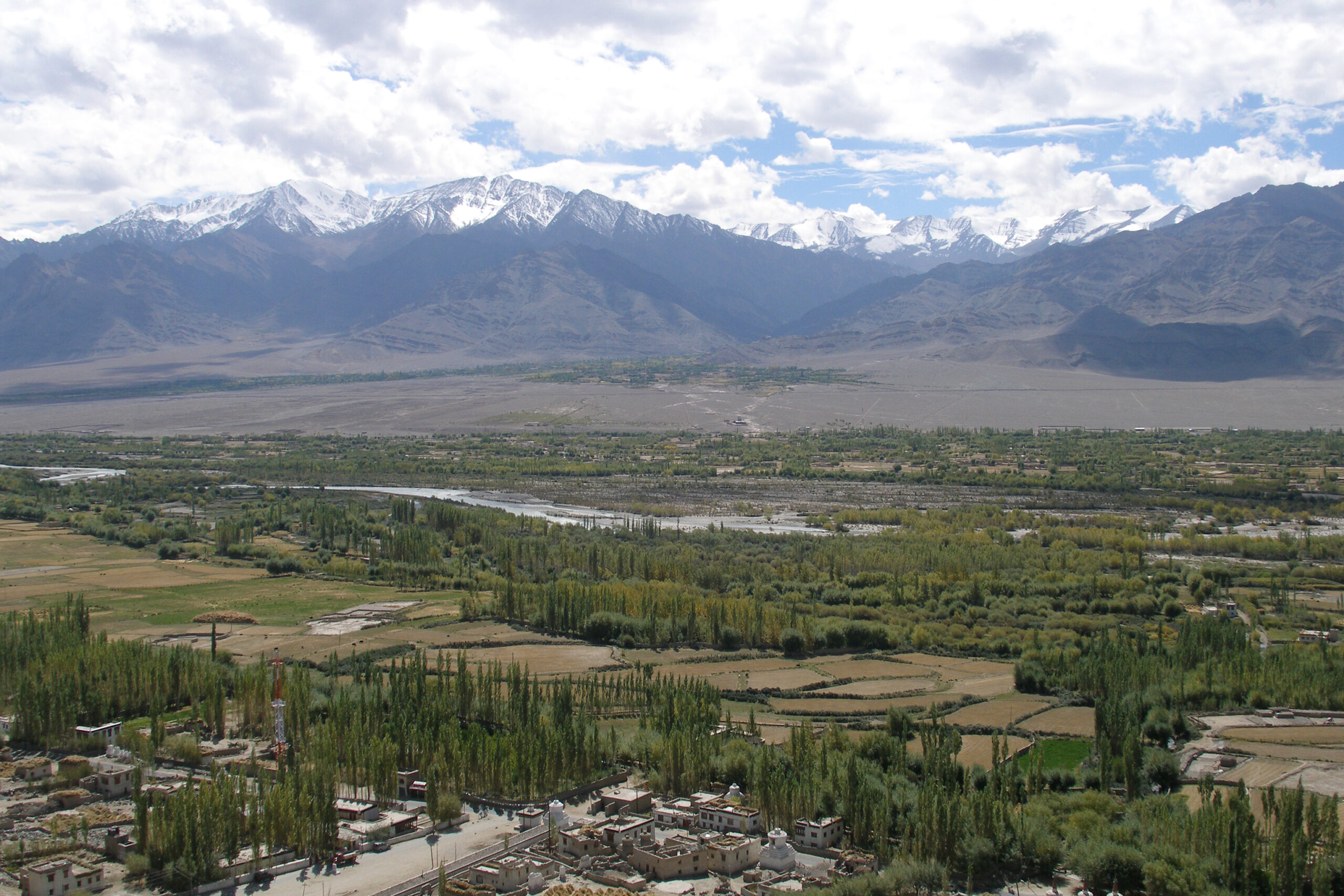A wide valley with green fields and scattered trees in the foreground, a river running through the middle, and snow-capped mountains under a partly cloudy sky in the background.