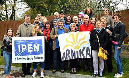 A group of people stand outdoors in front of a fence, holding two banners: one reads IPEN a toxics-free future and the other reads ARNIKA with a yellow sun-like design.