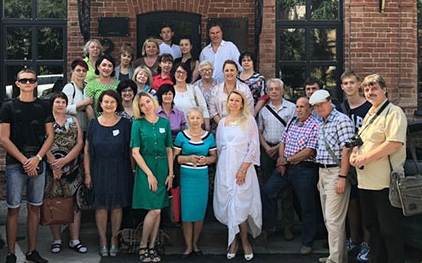 A group of about 30 people of various ages stands together posing for a photo in front of a building with red brick walls and dark doors. Most people are smiling and looking at the camera.