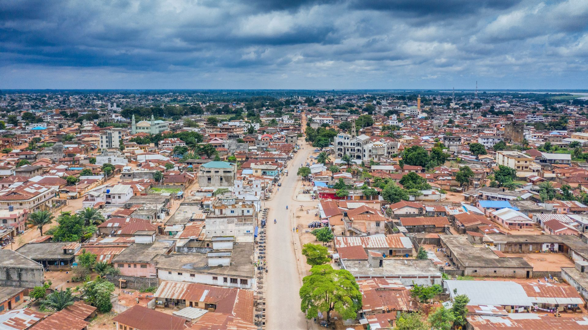 Aerial view of a city with a central road running through densely packed buildings, under a cloudy sky. The rooftops are mostly reddish-brown, and greenery is scattered throughout the area.