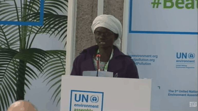 A woman wearing a white headwrap and purple jacket speaks at a podium with a microphone during the UN Environment Assembly. UN Environment logos and banners are visible in the background.