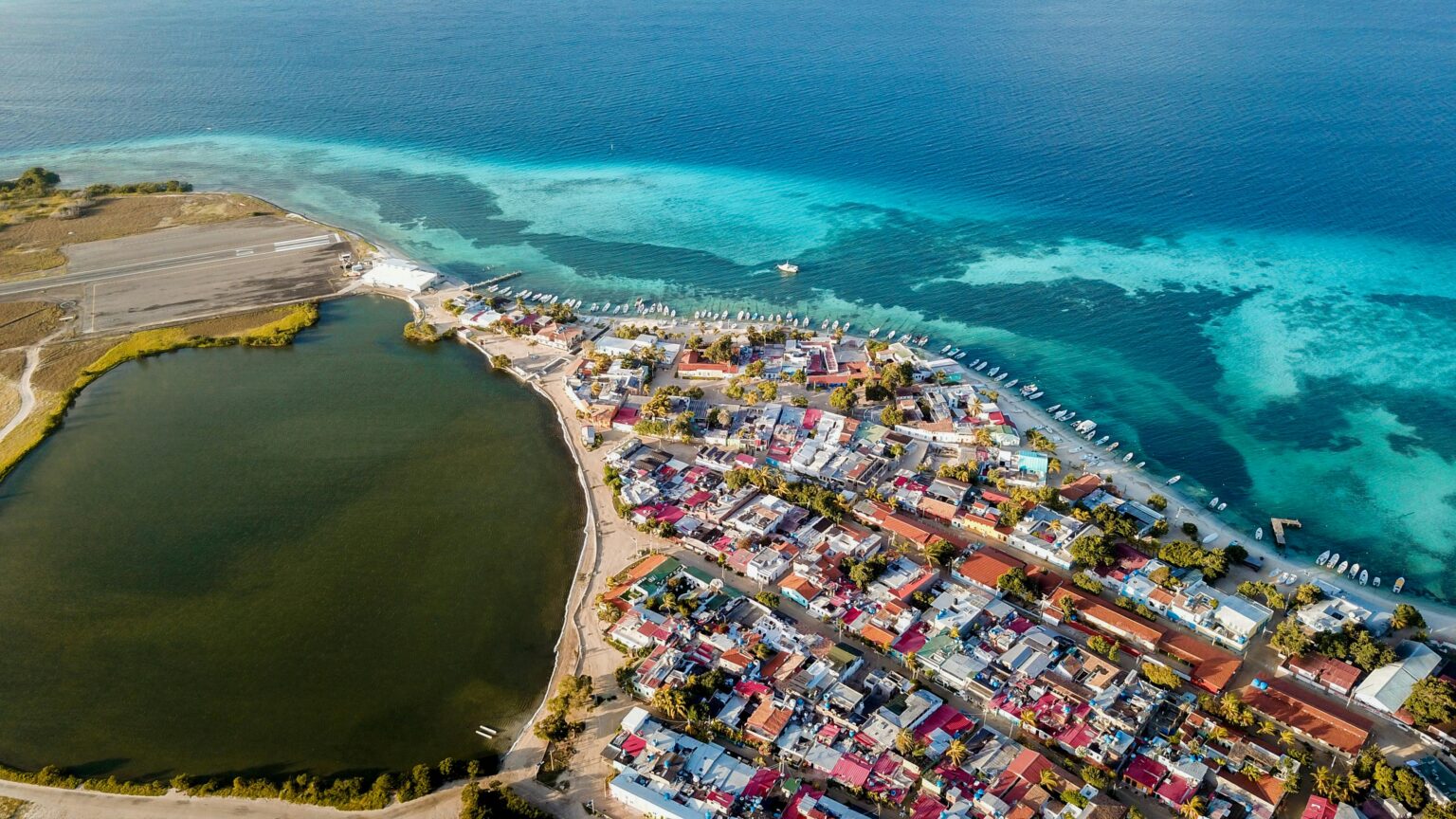 Aerial view of a coastal town with colorful buildings beside a lagoon and the ocean, featuring a dock with boats and an airstrip on the edge near the shoreline.