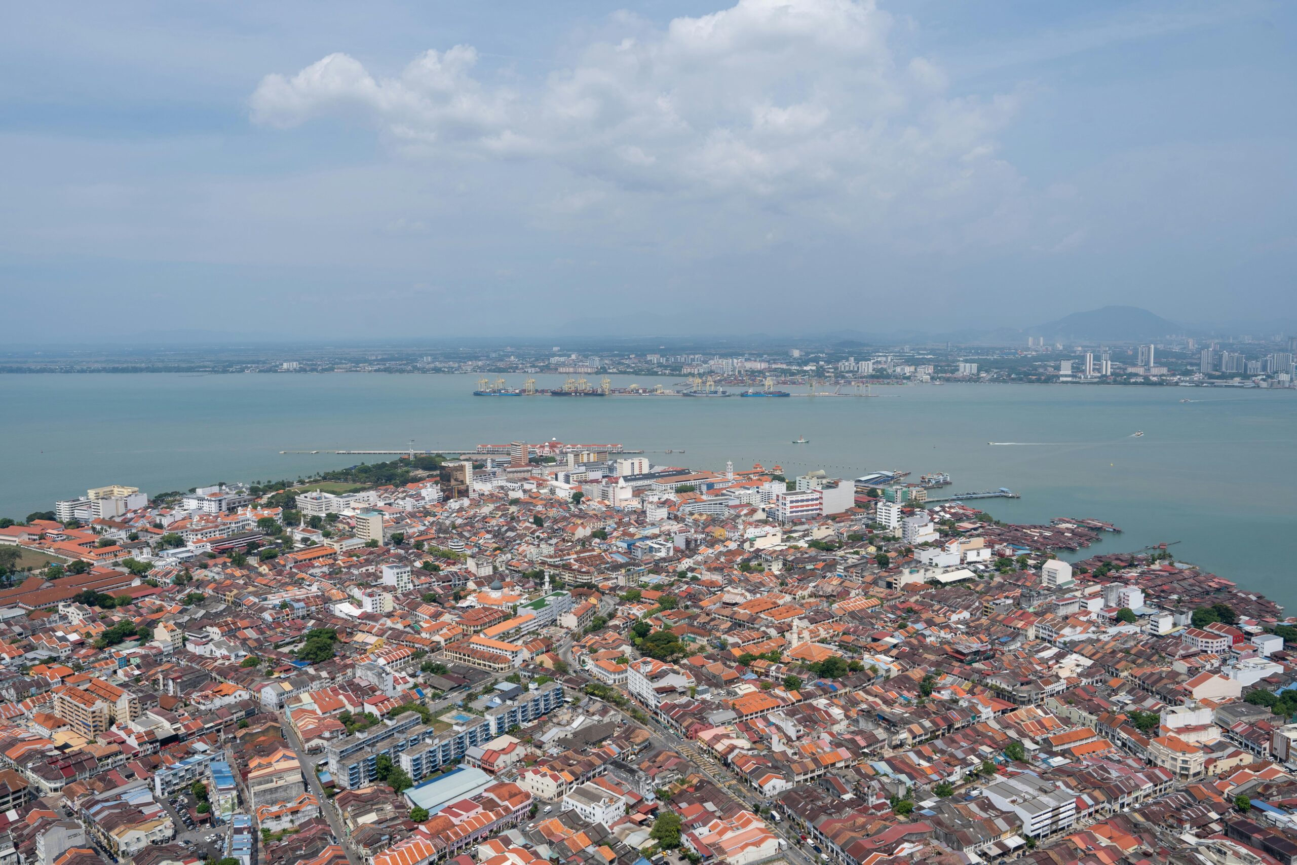Aerial view of a coastal city with densely packed buildings and red rooftops, bordered by a body of water with ships and a distant city skyline under a partly cloudy sky.