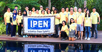 A group of people wearing yellow shirts stand outdoors next to a pool, holding a banner that reads IPEN a toxics-free future, with trees and greenery in the background.