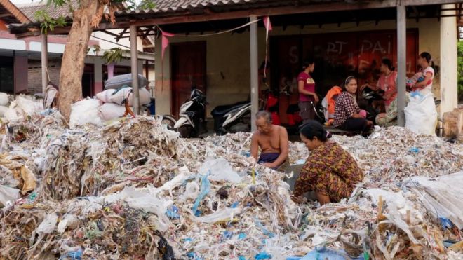 Several people sort through large piles of plastic and other waste outdoors near a building, with motorbikes parked under a roof in the background.