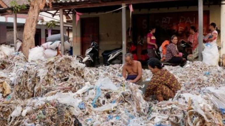 Several people sort through large piles of plastic and other waste in front of a building with motorbikes parked nearby. Some are seated amid the debris, while others stand or walk around.