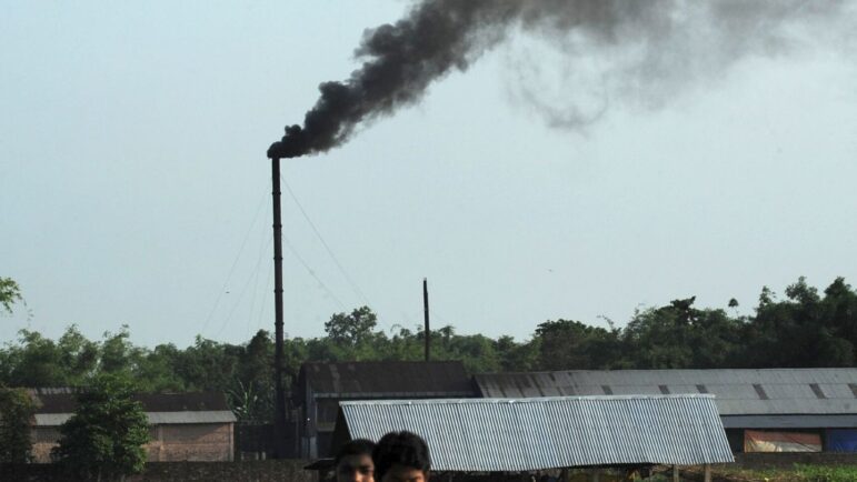 A tall industrial chimney emits thick black smoke above rooftops and trees under a clear sky, with two people partially visible in the foreground.