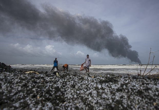 Three people walk along a rocky, debris-covered shoreline under a cloudy sky, with thick black smoke rising in the distance over the ocean.