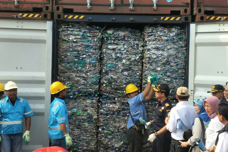 A group of people, some in uniforms and hard hats, inspect a shipping container packed tightly with compressed plastic waste. The container doors are open, and several people are taking notes or observing.