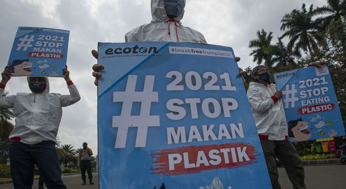 People wearing white hooded jackets and face masks hold blue signs that read #2021 STOP MAKAN PLASTIK and #2021 STOP EATING PLASTIC during an outdoor protest, with palm trees in the background.