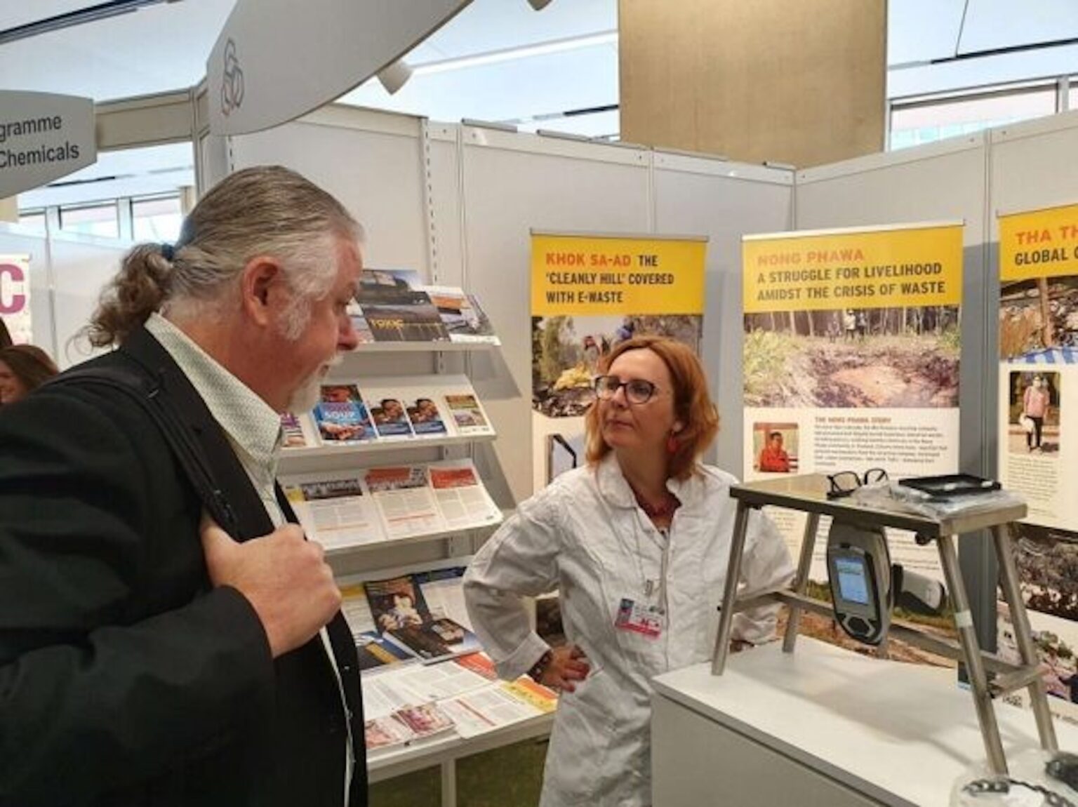 A man in a suit speaks with a woman in a white lab coat at an exhibition booth featuring informational posters and pamphlets about waste management and related challenges.