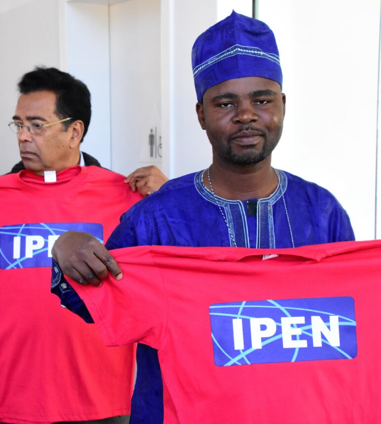 Two men hold up red T-shirts with a blue and white IPEN logo. The man in front wears a blue traditional outfit and cap, while the man behind him wears glasses and a black jacket.