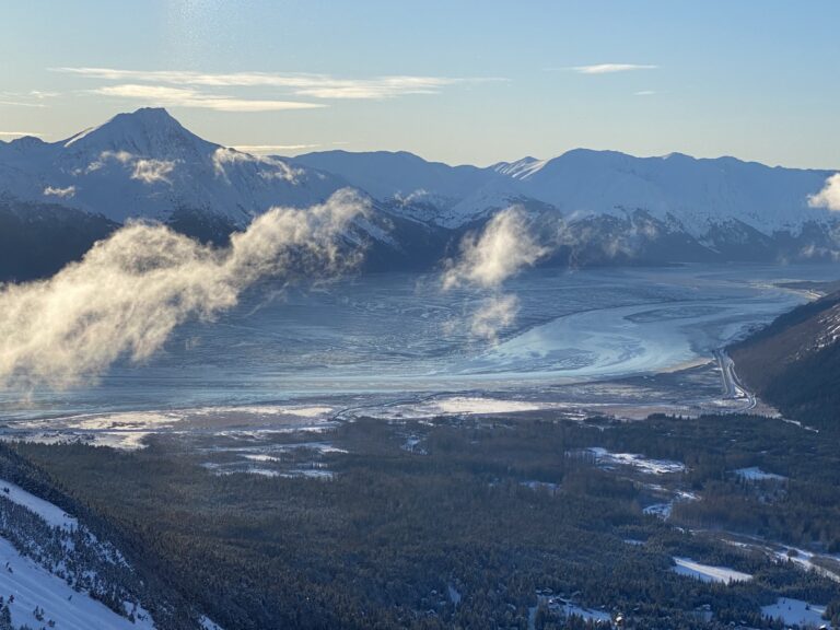 Snow-covered mountains and a winding, frozen river under a clear blue sky, with patches of thin clouds drifting above the landscape and forested areas in the valley below.
