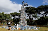 A sculpture of an oversized faucet appears to pour out a large pile of plastic bottles, spilling onto the grass, with a person standing nearby and trees in the background.