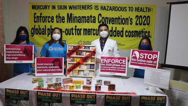Four people stand in front of a yellow banner about mercury hazards in skin whiteners, holding signs promoting the Minamata Convention and warning against mercury in cosmetics. Boxes of cosmetic products are displayed on the table.