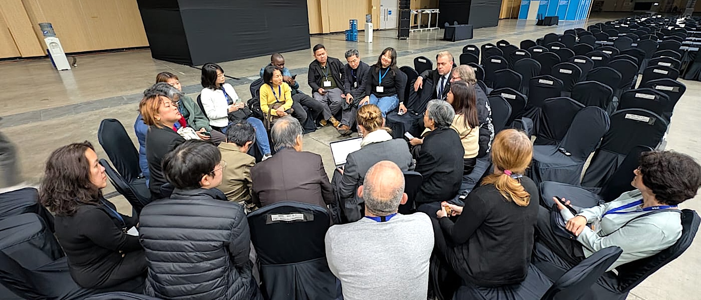 A group of people sit in a circle having a discussion in a large indoor hall. Rows of empty black chairs surround them, and some tables and equipment are visible in the background.