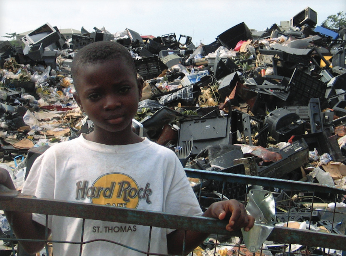 A young boy stands in front of a large pile of electronic waste and garbage, wearing a white Hard Rock Cafe St. Thomas t-shirt and resting his hand on a metal fence.