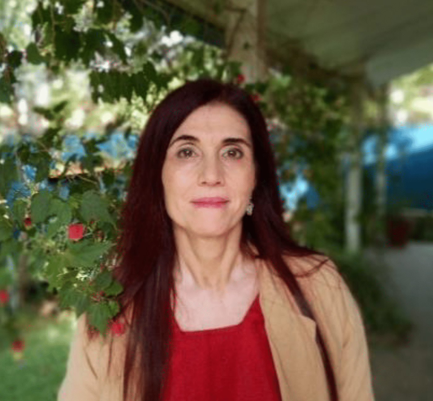 A woman with long dark hair and a neutral expression stands outdoors in front of green foliage, possibly attending an IPEN meeting. She is wearing a red top and beige jacket, with plants and a covered walkway visible behind her.