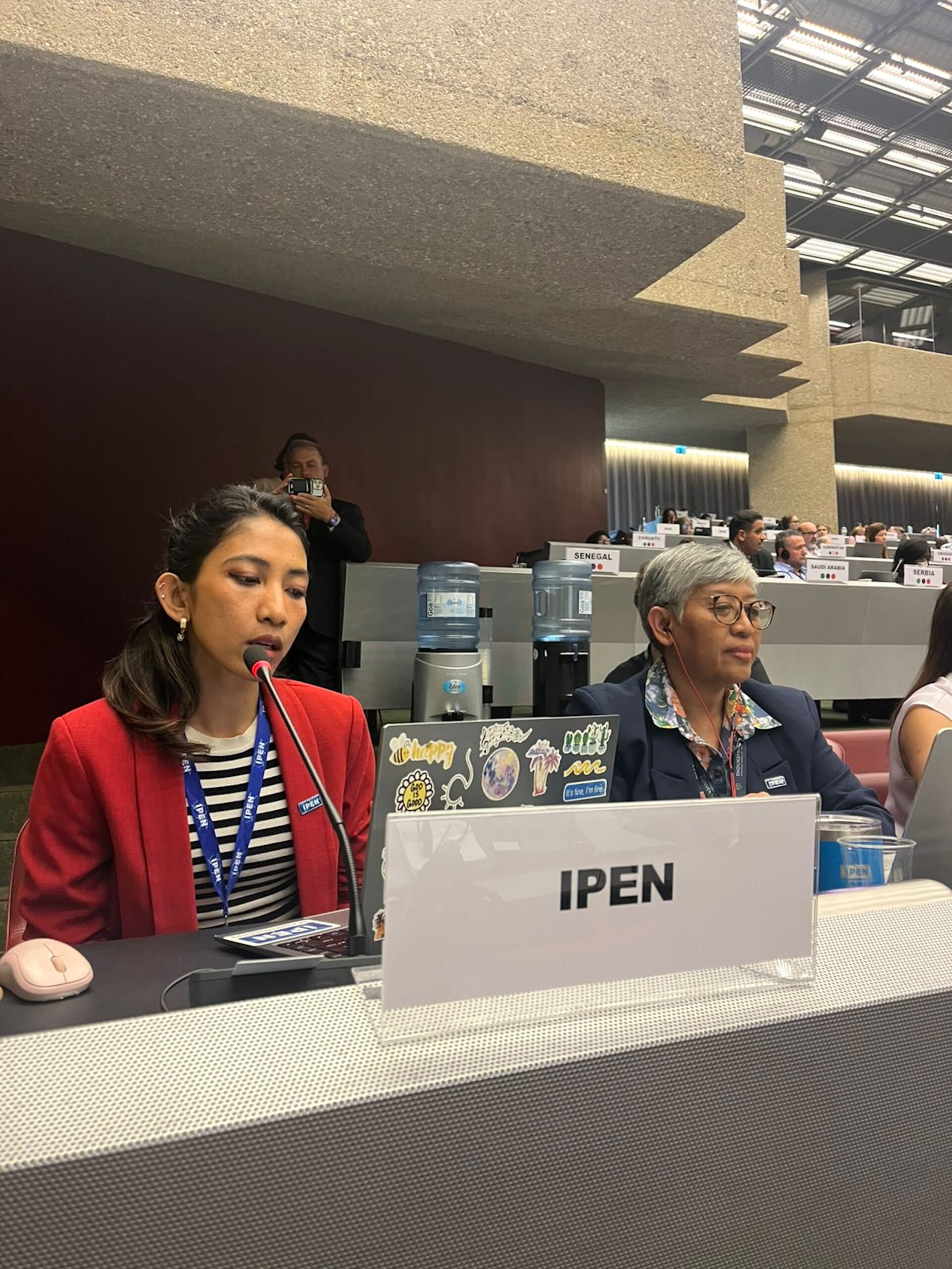 Two women sit at a conference table behind a sign labeled IPEN. One is speaking into a microphone, and both have laptops. Other attendees and a water dispenser are visible in the background.
