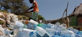 A person walks near a large pile of empty plastic bottles outdoors, with trees and simple structures visible in the background under a clear blue sky.