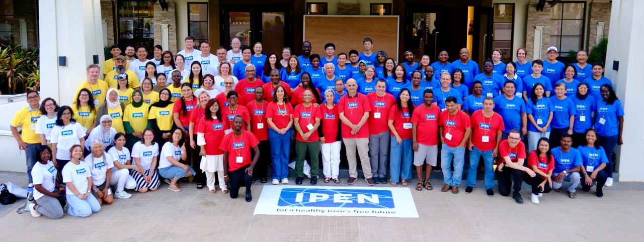 A large group of people pose for a photo outside a building, wearing red, blue, yellow, or white IPEN T-shirts. A banner on the ground reads IPEN for a healthy, toxics-free future.