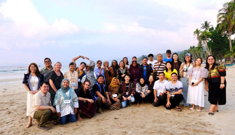 A group of about 30 people pose and smile for a photo on a sandy beach. Some are standing, others are crouching. The ocean and palm trees are visible in the background under a cloudy sky.