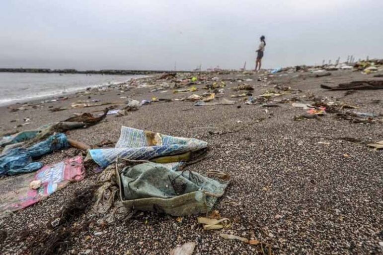 A beach covered with scattered plastic and other trash. A person stands near the shoreline in the background under a cloudy sky.