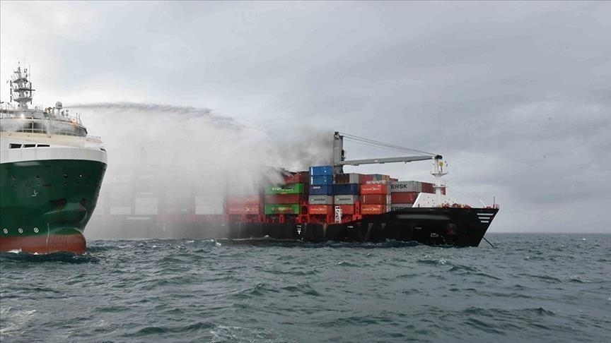 A vessel sprays water onto a cargo ship carrying stacked shipping containers, attempting to extinguish a fire. The ships are on choppy water under a cloudy sky.