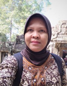 A woman wearing a brown patterned hijab and shirt takes a selfie outdoors during an IPEN meeting, standing in front of ancient stone structures with intricate carvings and trees in the background.