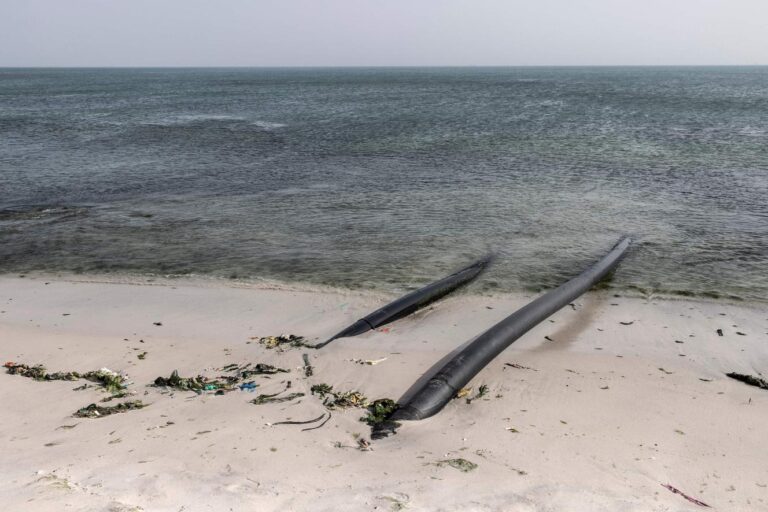 Two black pipes extend from the sandy shore into the ocean, with seaweed and debris scattered along the waterline under a cloudy sky.