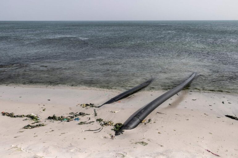 Two black pipes extend from the sandy shore into the ocean, with seaweed and debris scattered along the waterline under a cloudy sky.